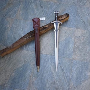 May include: A silver and black sword with a decorative handle and a brown leather sheath. The sword is lying on a piece of wood. The sheath is brown with a gold-colored tip.