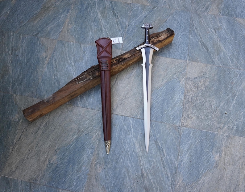 May include: A silver and black sword with a decorative handle and a brown leather sheath. The sword is lying on a piece of wood. The sheath is brown with a gold-colored tip.