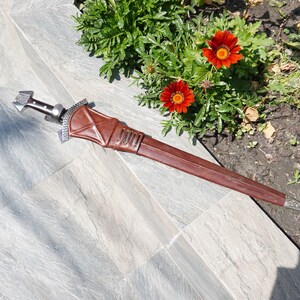 May include: A brown leather sheath with a silver-colored hilt and blade. The sheath is detailed with stitching and metal accents. The sword is lying on a gray tiled surface near red flowers.