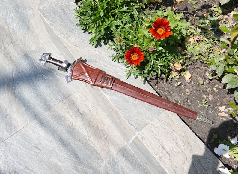 May include: A brown leather sheath with a silver-colored hilt and blade. The sheath is detailed with stitching and metal accents. The sword is lying on a gray tiled surface near red flowers.