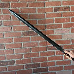 May include: A black sword with a wooden handle. The sword is held by a hand against a brick wall background.