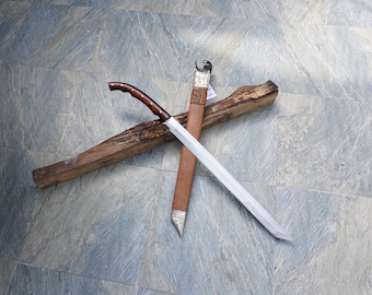 May include: A handmade sword with a wooden handle and a silver blade, resting on a piece of wood. The sword is sheathed in a brown leather scabbard with silver accents. The handle is dark brown with lighter brown accents.