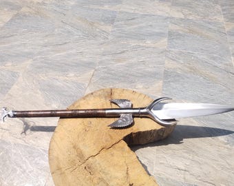 May include: A decorative spear with a silver blade and ornate details. The spear features a brown shaft with engraved patterns and silver wings near the blade. The spear is resting on a wooden surface.