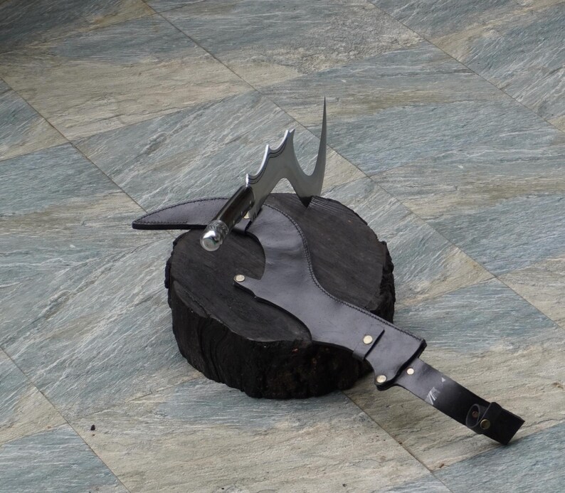 Puede incluir: Una funda de cuero negro con un hacha de lanzamiento de color plateado con una hoja curva. El hacha est&aacute; unida a la funda con una correa y una hebilla.