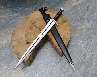 May include: A silver sword with a brown handle and a black sheath rests on a wooden surface. The sword has a double-edged blade with a central fuller. The sheath is made of black leather and has a silver tip.