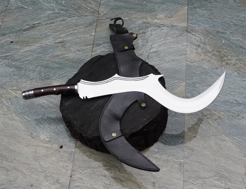 Puede incluir: Una hoja curva plateada y negra con un mango de madera y una funda de cuero. La hoja tiene un dise&ntilde;o decorativo y est&aacute; unida a la funda con una correa.