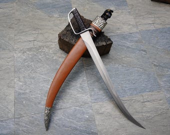 May include: A curved blade dagger with a brown leather sheath. The dagger features a silver handle and guard, and is resting on a piece of wood. The sheath has a silver tip.