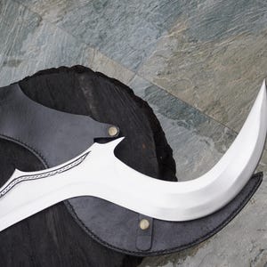 Puede incluir: Una hoja curva de color plateado con una funda de cuero negro. La hoja tiene un patr&oacute;n decorativo grabado.