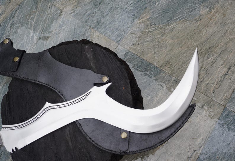 Puede incluir: Una hoja curva de color plateado con una funda de cuero negro. La hoja tiene un patr&oacute;n decorativo grabado.