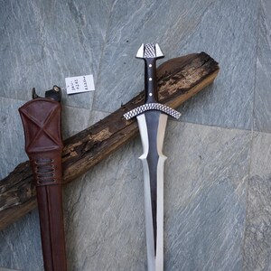 May include: A medieval-style sword with a silver blade and a black handle, resting on a wooden log. The sword has a decorative silver crossguard and pommel. A brown leather sheath is visible on the left side.
