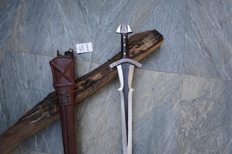 May include: A medieval-style sword with a silver blade and a black handle, resting on a wooden log. The sword has a decorative silver crossguard and pommel. A brown leather sheath is visible on the left side.