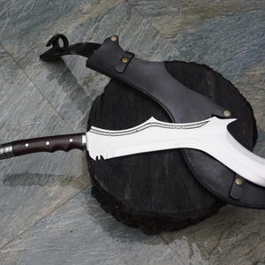 Puede incluir: Una hoja curva de color plateado con un mango de madera marr&oacute;n y una vaina de cuero negro. La hoja tiene un patr&oacute;n decorativo grabado.