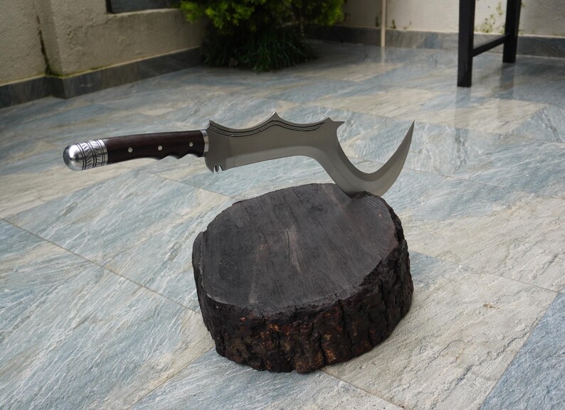 Puede incluir: Un cuchillo curvo plateado y marr&oacute;n con un mango decorativo, descansando sobre un toc&oacute;n de madera marr&oacute;n oscuro.