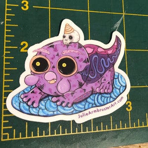 May include: A purple and blue cartoon creature with a skull on its head, sitting on a blue and white patterned surface. The creature has large yellow eyes and a pink mouth. The text "JulieArmbrusterArt.com" is printed on the surface.