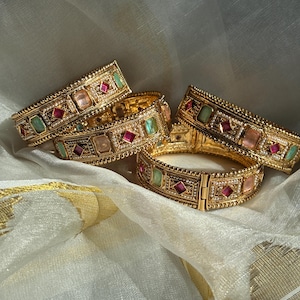 May include: Gold bangles with colourful gemstones. The bracelets feature square and diamond-shaped gems in shades of pink, green, and red, set in a detailed, ornate design. The bangles are displayed on a white and gold fabric.