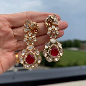 Puede incluir: Pendientes colgantes dorados con diseño de cabeza de elefante. Los pendientes tienen un diseño floral con piedras preciosas claras y rojas. Se muestran sobre un fondo borroso de vegetación y cielo nublado.