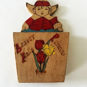 May include: Wooden wall hanging with a carved figure in a hat and red shirt above a wooden box. The box is painted with the words "Beaver Falls, Penna" and a bouquet of red and yellow tulips.