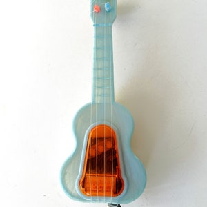 May include: A light blue plastic toy guitar with a clear orange sound box and a wind-up key. The guitar has a string design and colorful tuning pegs.