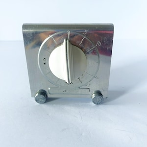 Puede incluir: Un temporizador de cocina metálico con base rectangular y dos pequeños pies cilíndricos. El dispositivo cuenta con un dial central de plástico blanco situado en una cara circular marcada con los números 5, 10, 15, 20 y 25, y un pequeño símbolo.