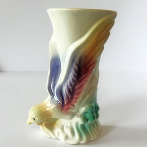 May include: A cream-colored ceramic vase featuring a sculpted bird in flight with blue and reddish-brown wing details. The base of the vase is decorated with green foliage accents, and the overall design has a glossy finish with a flared top rim.