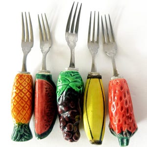 May include: Set of five decorative forks with fruit-shaped handles. The handles are designed as a pineapple, watermelon, grapes, banana, and strawberry. The forks have silver tines and colorful ceramic handles.