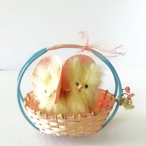 Puede incluir: Una cesta decorativa de Pascua con dos pollitos amarillos y esponjosos en su interior. Los pollitos tienen ojos negros y picos rojos. La cesta está hecha de material rosa claro tejido con un asa azul y una cinta rosa. Un pequeño adorno floral está unido al lateral.