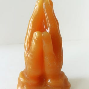Op de afbeelding: Oranje kaars in de vorm van biddende handen. De kaars heeft een lont en een rechthoekig etiket met tekst aan de basis. Op het etiket staat: "God grant me the serenity..." Deze kaars is een decoratief item.