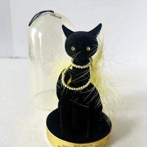 Puede incluir: Una figura de gato de terciopelo negro con un collar de perlas y detalles de plumas amarillas. El gato está sobre una base dorada con el texto "Max Factor Hypnotique Sophisti-Cat Perfume 1/8 fl. oz." Una cúpula de cristal transparente está detrás.