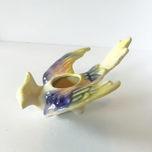 May include: A ceramic bird-shaped planter with a glossy finish. The bird is primarily cream-colored with blended accents of purple, blue, and yellow on its wings and tail. It features an open circular cavity on its back for small plants.