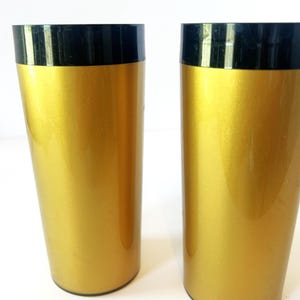 Op de afbeelding: Twee cilindrische bekers met een metallic gouden afwerking op het hoofdgedeelte en een contrasterende effen zwarte band rond de bovenrand. De vaten hebben een glad, reflecterend oppervlak en een recht ontwerp met een platte bodem.