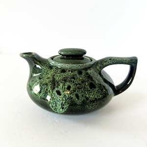 May include: A ceramic teapot featuring a glossy, mottled glaze in shades of dark forest green and vibrant lime green. The teapot has a rounded body, a short spout, a curved handle, and a matching lid with a small, circular knob on top.