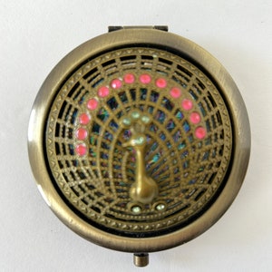 Pode incluir: Um espelho compacto redondo de cor dourada antiga com um design de pavão. As penas da cauda do pavão são iridescentes e o design é rodeado por um círculo de strass rosa.