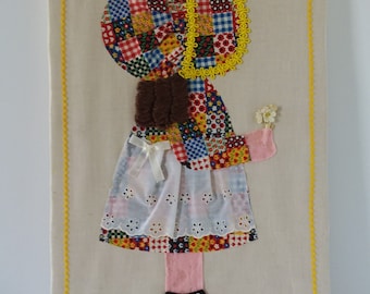 May include: A vintage patchwork fabric doll with a white bonnet and a white dress with lace trim. The doll is holding a small white flower in its hand. The doll is mounted on a beige fabric background with a yellow stitched border.