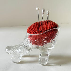 May include: A clear glass shoe-shaped pin cushion with a red velvet cushion. Three pearl-headed pins are inserted into the cushion. The glass has a cut-glass pattern, and the shoe has three small feet.
