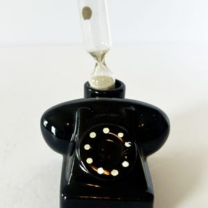 May include: A glossy black ceramic telephone-shaped object featuring a circular rotary dial with white dots. A clear glass hourglass containing light-colored sand is inserted into the top of the telephone, extending vertically from the base.