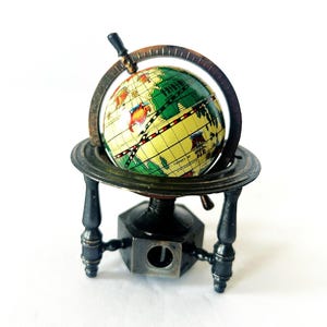 Puede incluir: Un sacapuntas de globo en miniatura. El globo presenta un diseño de mapa amarillo y verde, enmarcado en un marco y soporte de metal oscuro. La base tiene una abertura para sacapuntas. Un accesorio de escritorio decorativo y funcional.