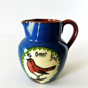 May include: A small blue ceramic pitcher with a brown handle and rim. The front features a circular cream-colored medallion with a crackled glaze, displaying a painted robin perched on a branch and the black handwritten text To Greet You.