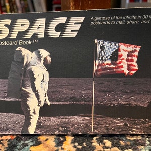 May include: A postcard book titled "SPACE" with a black background and a photo of an astronaut standing on the moon with an American flag planted in the lunar surface. The text "A Postcard Book™" is below the title.