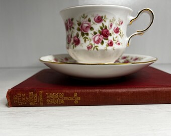May include: A red book titled "The Little Book of Modern Verse" by  E.V. Rieu sits on a white surface with a white teacup and saucer with a pink floral pattern on top of it.