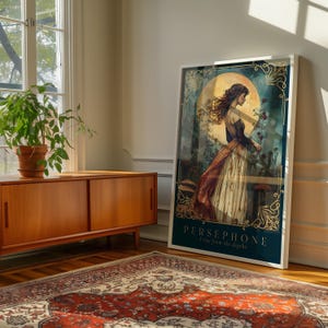 Op de afbeelding: Een ingelijste kunstdruk met een vrouw in een vloeiende jurk, tegen een achtergrond van een grote maan en bloemelementen. Het kunstwerk is ingelijst in een witte rand en bevat de tekst "PERSEPHONE I rise from the depths."