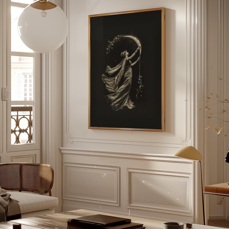 May include: A framed art print featuring a woman reaching for a crescent moon, with stars and flowing fabric. The artwork is in grayscale with a black background and a gold frame, creating a vintage aesthetic. The print is displayed on a white wall.
