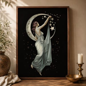May include: A framed wall art print featuring a figure in a flowing light-colored gown reaching toward a crescent moon and stars against a black background. The figure is positioned centrally, with the fabric of the gown draping downward.