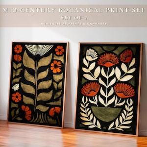 May include: Two framed botanical art prints. The prints feature stylized floral designs in orange, gold, and white against a black background. The frames are a warm, light brown color. The text at the top reads "MID CENTURY BOTANICAL PRINT SET" and "SET OF 2".