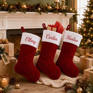 Puede incluir: Tres calcetines navideños de terciopelo rojo intenso con puños de piel sintética blanca. Cada calcetín tiene un nombre bordado en letra roja: Ellery, Corbin y Harlow. El calcetín central contiene una pequeña caja de regalo envuelta en papel beige.