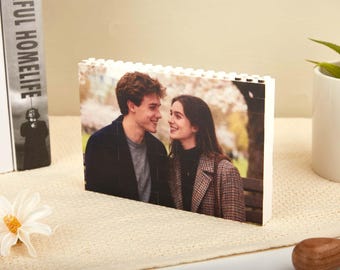 May include: A rectangular photo printed on white blocks, featuring a couple looking at each other. The man wears a dark jacket over a gray shirt, and the woman wears a patterned coat. The photo is displayed on a beige surface.