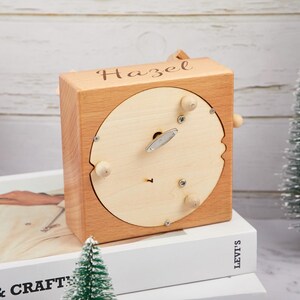 Puede incluir: Una caja de m&uacute;sica de madera con una inscripci&oacute;n personalizada que dice "Hazel". La caja de m&uacute;sica est&aacute; hecha de madera marr&oacute;n claro y tiene una abertura circular en la parte superior. La caja de m&uacute;sica est&aacute; sentada sobre una pila de libros.