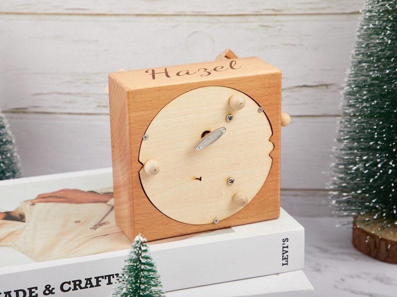 Puede incluir: Una caja de m&uacute;sica de madera con una inscripci&oacute;n personalizada que dice "Hazel". La caja de m&uacute;sica est&aacute; hecha de madera marr&oacute;n claro y tiene una abertura circular en la parte superior. La caja de m&uacute;sica est&aacute; sentada sobre una pila de libros.
