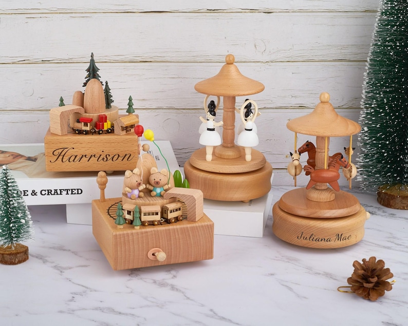 Puede incluir: Tres cajas de m&uacute;sica de madera con nombres personalizados. La primera caja de m&uacute;sica presenta un tren y &aacute;rboles, la segunda caja de m&uacute;sica presenta un carrusel con caballos y la tercera caja de m&uacute;sica presenta un carrusel con bailarinas. Las cajas de m&uacute;sica est&aacute;n hechas de madera y tienen un acabado natural.