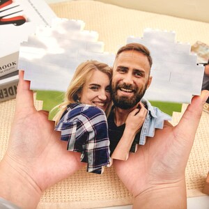 Könnte beinhalten: Ein herzförmiges Foto-Puzzle mit einem lächelnden Paar. Das Puzzle besteht aus ineinandergreifenden Teilen, auf denen das Bild des Paares abgebildet ist. Der Hintergrund zeigt eine grüne Wiese und einen bewölkten Himmel. Das Puzzle wird in zwei Händen gehalten.