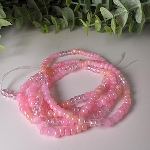 May include: A collection of pink and clear waist beads. The beads are in varying shades of pink, from light to darker hues, and are strung together. The beads are small and round, with a clear, transparent section interspersed throughout.